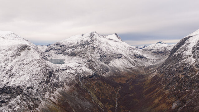 Majestic mountain valley in autumn, surrounded by snow-capped peaks and rugged slopes. A winding river flows through the valley floor with patches of green fields beneath a cloudy, dramatic sky.
