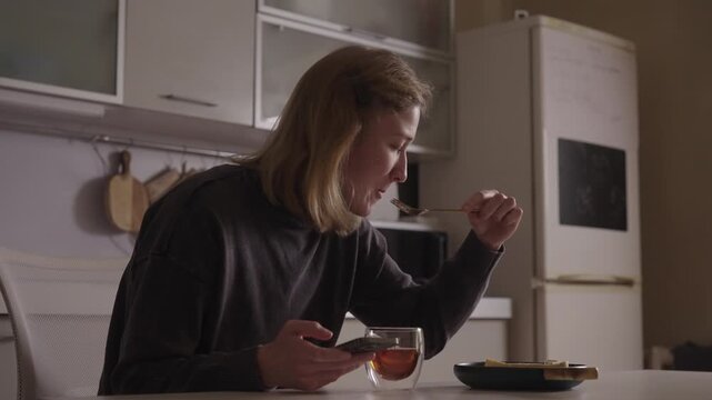 Young woman living with cerebral palsy focusing on smartphone screen while enjoying dessert and a warm cup of tea at kitchen table, representing independence and normalcy in daily home life