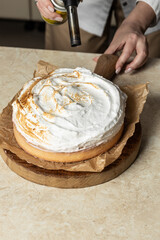 A chef uses a culinary torch to caramelize the meringue on a round cake. The dessert sits on a wooden board lined with parchment paper, highlighting the golden-brown peaks of the toasted meringue.