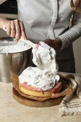 A chef spreads fluffy white meringue over a cake with a spatula. The cake, topped with pink filling, sits on a wooden board lined with parchment paper. A close-up of the dessert decorating process.