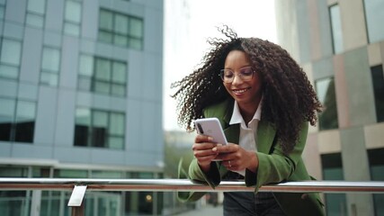 Young African American business woman in trendy outfit and glasses using mobile phone on corporate urban district. Creative female professional in the city 