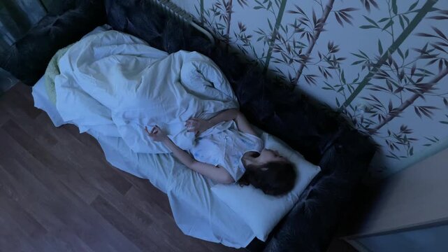 Dnipro, Ukraine - January 4, 2021: Cosplayer girl in image of Nancy Thompson from horror film Nightmare on Elm Street lying in bed suddenly waking up from nightmare in dark bedroom. High angle shot