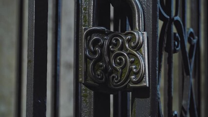 Close-up video of an ornate padlock on a wrought iron gate, captured at an angle highlighting intricate details and textures in low light. - Powered by Adobe