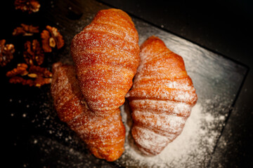 Three freshly baked golden croissants, heavily dusted with powdered sugar, are beautifully presented with walnuts and berries on a moody, dark slate board, perfect for breakfast or dessert menus