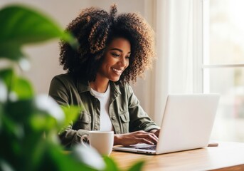Smiling young black woman working on laptop at home with coffee and plant
