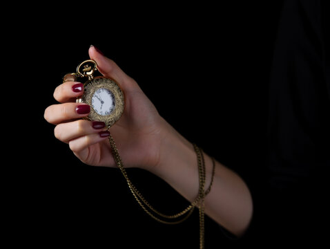 A woman's hand with elegant burgundy nails holds a beautiful vintage golden pocket watch with an ornate design and chain, set against a dramatic black background. Symbolizing time, history, and precis