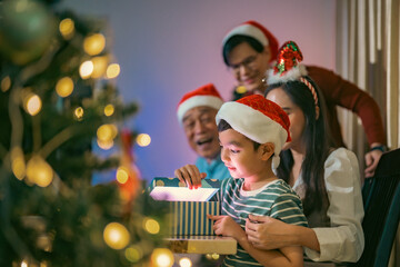 Excited Asian boy opening a Christmas gift with family cheering beside him, creating a heartwarming...