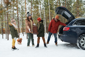 Woman handing picnic basket to teenager boy while man unloading car and child standing nearby in snowy forest