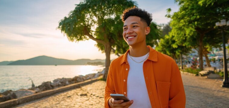 The young man strolling along waterfront promenade at sunset holding smartphone and smiling - Powered by Adobe