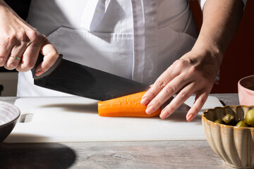 Chef in white apron slices a carrot on a white cutting board with a large kitchen knife. Close-up of hands at work, with a bowl of olives visible in the background. Focus on the food preparation 