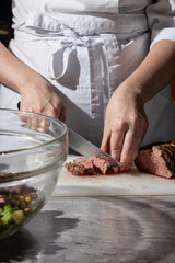Chef in a white apron carefully slices a medium-rare steak on a white cutting board. A glass bowl...