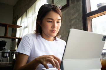 A Focused Asian Thai Young Woman Intently Working on a Tablet in a Cozy Cafe Environment.