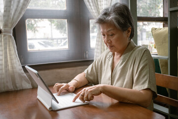 Asian elderly woman types on a laptop at a cozy wooden table, with natural light enhancing her focus and enjoyment
