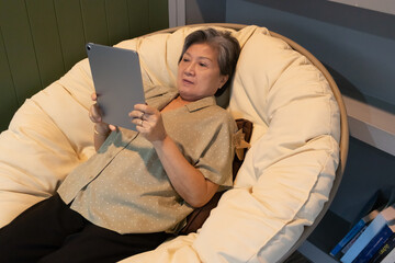 Asian elder woman Enjoying a Relaxing Moment Reading on a Comfortable Tablet while Nestled in a Cozy Bean Bag Chair