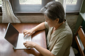 Asian elderly woman types on a laptop at a cozy wooden table, with natural light enhancing her focus and enjoyment