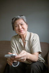 A Joyful and Smiling Asian Older Woman Holding a mobile phone in a Cozy and Comfortable Sofa.