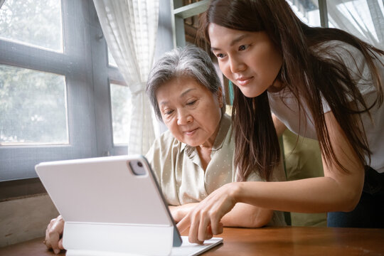 Asian Thai woman patiently helps an elderly lady use a tablet, showcasing the beauty of connection and learning at cozy house.