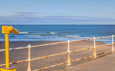 A pier with a telescope in the foreground and a ship on the horizon.