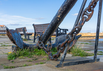 An ancient weathered anchor is on a quay with weeds growing around it.