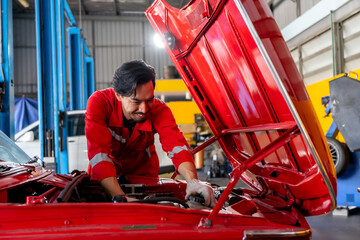happy and skilled Asian male mechanic in a red uniform smiles while working intently on the engine of a bright red classic car with the hood open