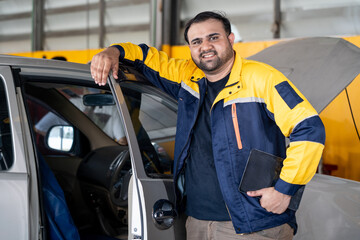 A friendly and confident Indian male mechanic or service manager in a yellow and blue uniform leans against a car door with the hood open, holding a tablet. His genuine smile