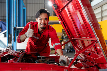 happy and skilled Asian male mechanic in a red uniform smiles and show thumbs-up while working intently on the engine of a bright red classic car with the hood open in car service auto repair shop
