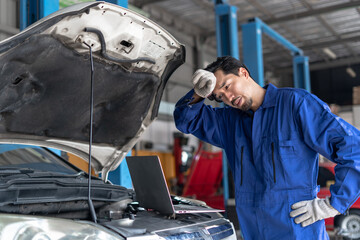 A frustrated Asian mechanic wipes sweat while facing a complex computer diagnostics issue with an open-hood vehicle. Japanese mechanic man in blue uniform Stressed car mechanic in repair garage
