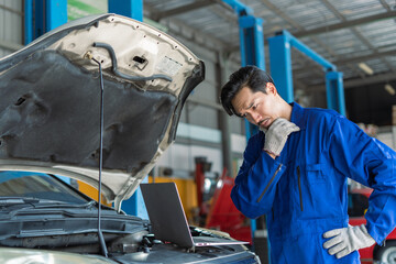 thoughtful Asian male mechanic stroking his chin in deep concentration working with laptop on the engine bay troubleshooting a complex issue found through computer diagnostics in a professional garage