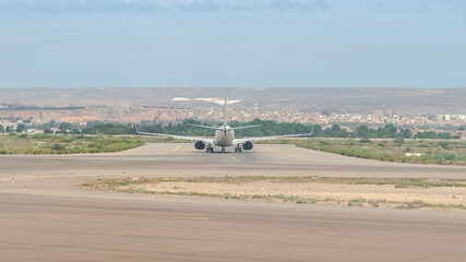 Fototapeta premium Rear view of a commercial airplane taxiing on the runway preparing for takeoff, with forest trees, old city buildings and a distant mountain under a clear blue sky.