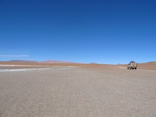 A vehicle moves across the expansive Altiplano desert in Bolivia, heading towards Uyuni. The vast, empty landscape and blue sky create a dramatic and remote wilderness scene.