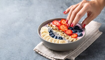 hand with red nail polish touching smoothie bowl with banana strawberry blueberry coconut and granola on gray background
