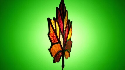 Stained glass feather in warm autumn colors of red, orange, and yellow glowing against a vibrant green background - Powered by Adobe