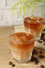 Tasty iced coffee with milk in glasses and beans on wooden table against light textured background, closeup