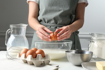 Making batter (liquid dough). Woman adding eggs into bowl at white table indoors, closeup
