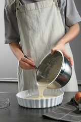 Woman pouring liquid dough from bowl into baking dish at grey table in kitchen, closeup