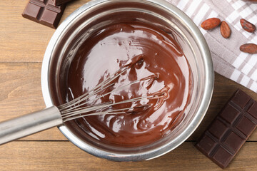 Melted chocolate with whisk in bowl, beans and pieces on wooden table, flat lay
