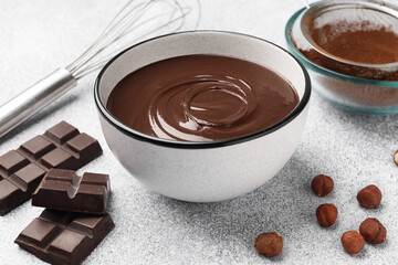Melted chocolate in bowl, pieces, nuts, cocoa powder and whisk on light grey table, closeup