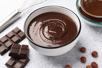 Melted chocolate in bowl, pieces, nuts, cocoa powder and whisk on light grey table, closeup