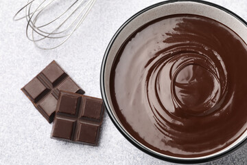 Melted chocolate in bowl, pieces and whisk on light grey table, flat lay