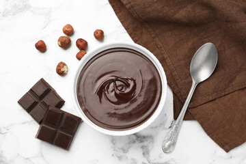 Melted chocolate in bowl, pieces, nuts and spoon on white marble table, flat lay