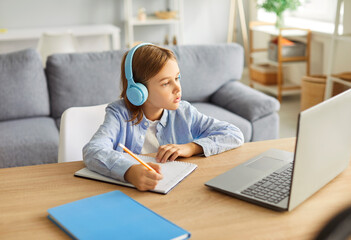 Focused preteen child wearing headphones sitting at table in front of laptop and listening to lecture. Preteen kid writing notes in notebook with pencil, studying online. Distance learning concept.