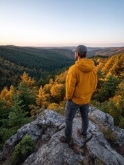Naklejka premium A man in a yellow jacket stands on a rock, gazing at a vast forest landscape during autumn. The scene captures tranquility and adventure in nature.