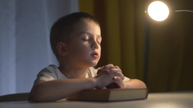 Young boy is praying. Religious leader prays to a child. Lifestyle book of prayers on a table in the evening. A boy is offering a prayer.