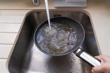 Hand rinses oily frying pan under faucet in stainless sink, unsafe contact of water with hot oil and visible splashes