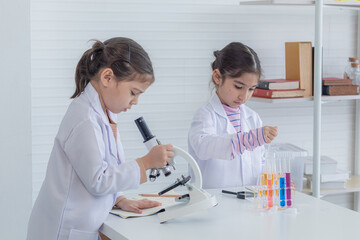 Cheerful elementary student girls wearing safety eyeglasses using magnifier glasses and microscope in lab coat uniform, sibling kids having fun with experiment test in science class future scientist