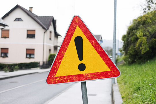 Warning road sign with exclamation mark near houses. Attention symbol on street in residential area. Safety and traffic caution concept with triangle sign on roadside and suburban background.