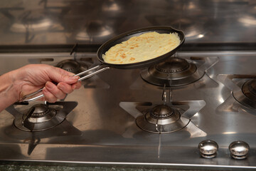 Woman holding frying pan with freshly cooked crepe over stainless steel gas stove in home kitchen
