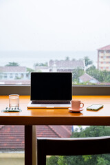 Cozy remote workstation with laptop computer, coffee mug and smartphone on wooden desk by window with rain outside.