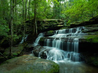 The waterfall looks smooth gentle, multi-tiered, beautiful flowing over mossy rocks in the lush green forest behind, dense lush, with tall trees dense bushes, Tourist attractions in Thailand