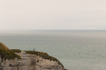 A lone figure standing on the edge of a white cliff overlooking the calm sea at Étretat, Normandy. Peaceful coastal landscape evoking solitude and vastness.